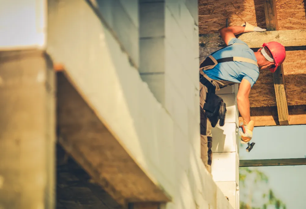 Construction worker wearing a red helmet and blue shirt installing exterior materials on a building addition, representing Restorations Unlimited’s seamless and efficient commercial expansion services in Maine.