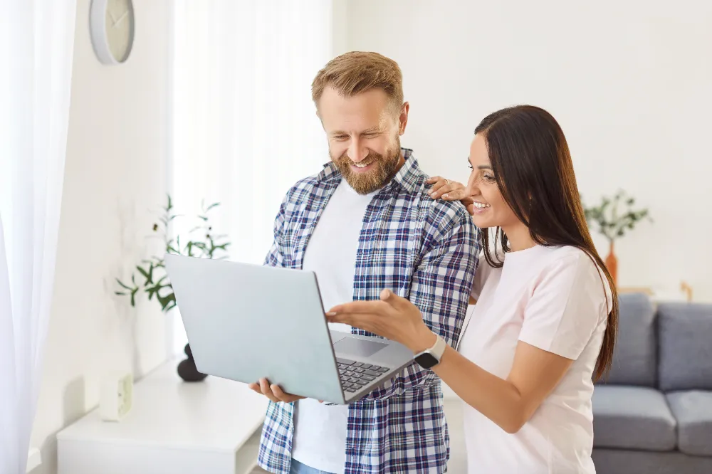 A smiling couple reviewing renovation plans together on a laptop in their living room, representing the clarity and organization provided by the Restorations Unlimited Project Build Plan.