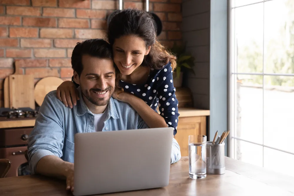 Smiling couple reviewing renovation plans together on a laptop in their kitchen, symbolizing how Restorations Unlimited’s Project Build Plan creates reliable timelines and eliminates costly surprises before construction begins.