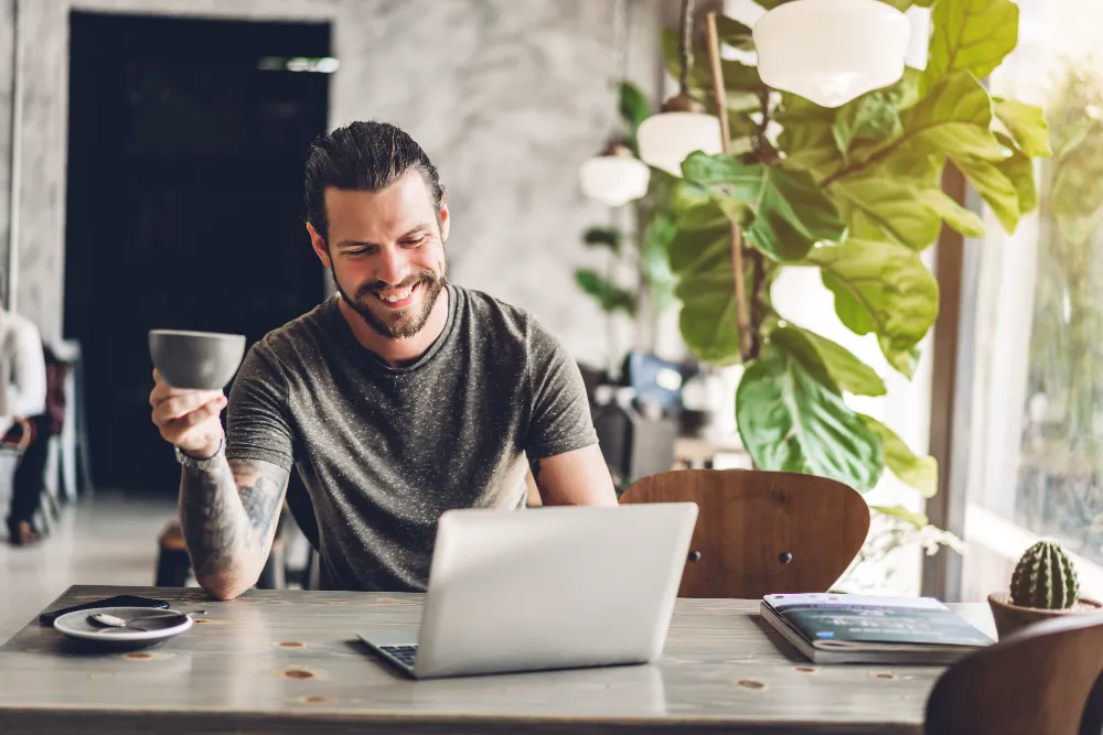 Man smiling while working on a laptop with a coffee mug in hand, symbolizing the thoughtful planning, design functionality, and stress-free project management provided by Restorations Unlimited.