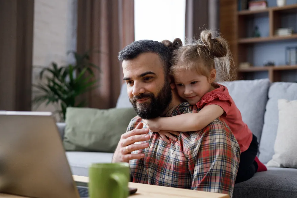 Father working on a laptop at home while his daughter hugs him from behind, symbolizing how Restorations Unlimited’s Project Build Plan creates peace of mind and smoother coordination for families during renovations.